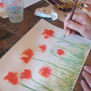 Gros plan sur une main peignant des fleurs rouges à l’aquarelle sur papier lors de l’atelier découverte, espace d’exploration créative et florale. 