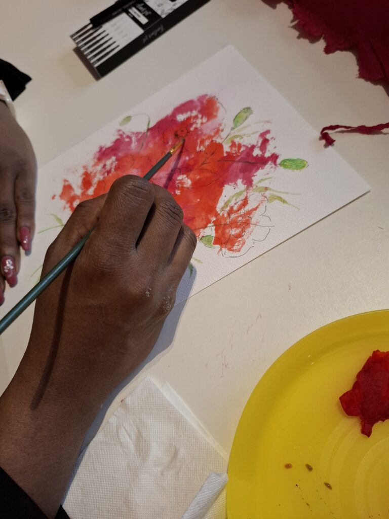 Atelier découverte aquarelle florale : main d’une participante peignant une fleur rouge sur carte blanche, animation créative et florale à Liège.