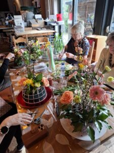 « Participants assis autour d’une grande table en intérieur, ajoutant les derniers détails à leurs compositions florales lors de l’étape “Finition et partage” de l’atelier. 